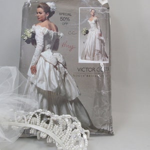 May include: A vintage wedding dress pattern by Victor Costa, featuring a bridal gown with long sleeves and a full skirt. The image includes a white veil and a pearl-adorned tiara, suggesting a bridal theme. The pattern is marked "Special 50% Off".