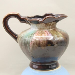 May include: A ceramic pitcher featuring a glossy dark brown glaze with flowing, drip-style accents in shades of cream, soft pink, and muted green. The vessel has a curved handle, a scalloped rim, and a rounded body that tapers to a circular base.