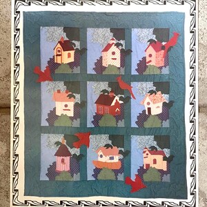 May include: A square fabric quilt featuring a grid of nine blocks, each displaying a different birdhouse design in shades of red, pink, and cream. The quilt is accented with dark fabric birds and a decorative black and white geometric border.