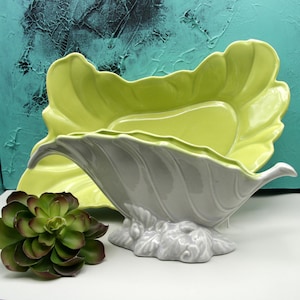 May include: Two ceramic leaf-shaped serving dishes. One is light green, the other is gray. The gray dish has a textured, curved design and sits on a decorative base. A green and brown succulent is in the foreground.