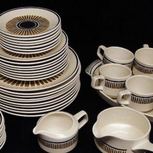 May include: A dinnerware set featuring off-white ceramic plates, cups, and pitchers with a dark brown sunburst or vertical line pattern. The collection includes stacked plates of varying sizes, multiple handled cups, and two pitchers on a black background.