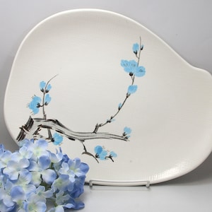May include: White ceramic serving platter with a unique, asymmetrical shape. The platter is decorated with a branch of blue flowers and a black stem. The platter is on a white stand.