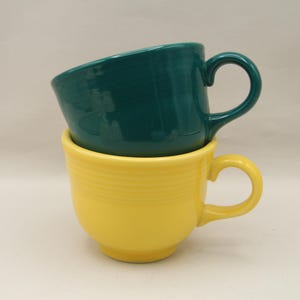 May include: Two ceramic mugs stacked on top of each other. The top mug is teal, and the bottom mug is yellow. Both mugs have a rounded shape and a curved handle. The yellow mug has horizontal ridges around the body.