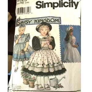 May include: Simplicity sewing pattern featuring a "Daisy Kingdom" design. The pattern includes illustrations of children's dresses with floral and lace details. The package is white with black text and a barcode.