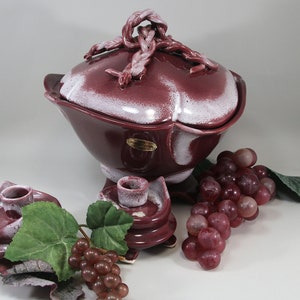 May include: A burgundy ceramic serving dish with a matching lid and two candle holders. The dish and candle holders have a white, textured trim. A bunch of grapes and green leaves are arranged near the dish.