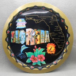 May include: Round black tray with a gold rim featuring the word "FLORIDA" in colorful letters. The tray includes images of a parrot, a marlin, hibiscus flowers, and a sunset. A map of Florida with city names is also displayed.