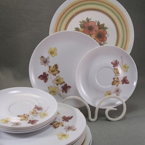 May include: A collection of white ceramic plates and saucers with autumn leaf and floral designs. The plates vary in size, with some featuring a floral pattern and others with a leaf motif in shades of brown, yellow, and orange.