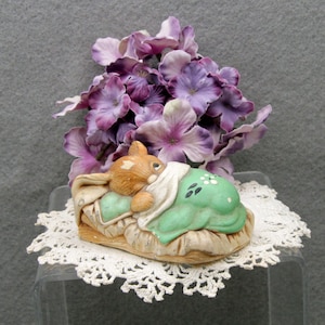 May include: A ceramic figurine of a sleeping bunny on a green pillow with white polka dots, covered by a white blanket. The bunny is light brown with a white patch. The figurine rests on a beige base, with purple flowers in the background.