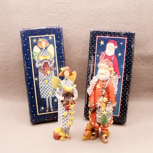 May include: Two decorative figurines and their matching rectangular boxes. One figurine is an angel in a blue and white checkered dress with yellow stars. The other is a Santa Claus in a red coat with a sack of toys and a small teddy bear.
