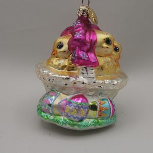 May include: An Easter ornament featuring three golden chicks nestled in a nest. The ornament has a pink and purple draped fabric and is decorated with colorful Easter eggs. The ornament is approximately 4 inches tall.