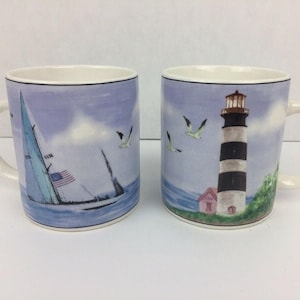 May include: Two ceramic mugs with coastal designs. One mug features a sailboat with an American flag, the other a black and white striped lighthouse. Both mugs have a blue and white color scheme with seagulls and a light blue sky.