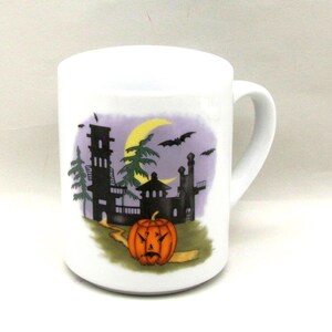 May include: A white ceramic mug featuring a Halloween-themed graphic. The illustration depicts a black silhouette of a haunted castle, a yellow crescent moon, flying bats, and a carved orange jack-o'-lantern pumpkin sitting on a path.