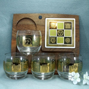 May include: Set of four clear glass tumblers with gold and green decorative bands featuring sun and lyre designs. The glasses sit on a wooden serving tray with a matching tile coaster. The coaster has a gold and green geometric pattern.
