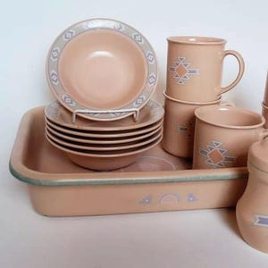 May include: A peach-colored ceramic dinnerware set featuring plates, bowls, mugs, and a serving dish. The plates and bowls have a decorative border with geometric shapes. The mugs and serving dish have a geometric design. The set is arranged on a white surface.