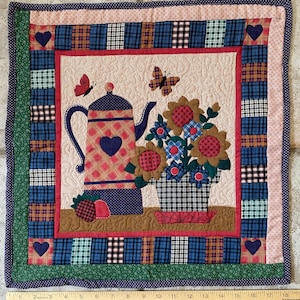 May include: A quilted wall hanging featuring a teapot and a vase of flowers. The teapot has a heart-shaped design, and the flowers are red, blue, and yellow. The border is a patchwork of plaid and polka dots, with heart accents.
