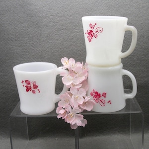 May include: Three white ceramic mugs with red floral designs are stacked on a clear surface. The mugs have handles and are decorated with a red floral pattern. Pink flowers are placed in front of the mugs.