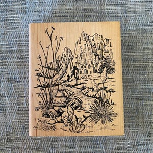 May include: A rectangular wooden stamp featuring a black and white desert landscape scene. The design includes mountains, cacti, pottery, and desert plants. The stamp is made of light-colored wood.