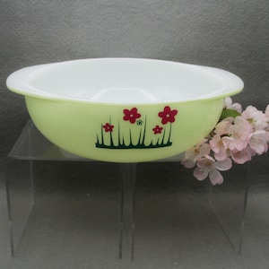 May include: A round, light green bowl with a white interior and integrated side handles. The exterior features a dark green grass graphic with several red flowers and one small green flower printed on the side of the bowl.