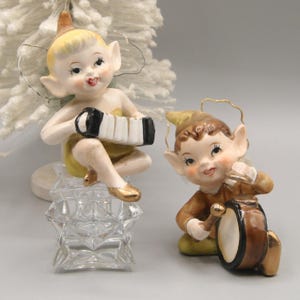 May include: Two ceramic elf figurines. One elf plays an accordion, the other a drum. The elves have blonde and brown hair, rosy cheeks, and are dressed in yellow and brown outfits. The accordion is black and white.