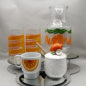 May include: A vintage orange-themed glassware set. Includes two tall glasses with "ORANGE" text, a pitcher with orange and green designs, a white mug with an orange slice, and a white ceramic jar with a lemon lid. A wooden stirring stick is also present.