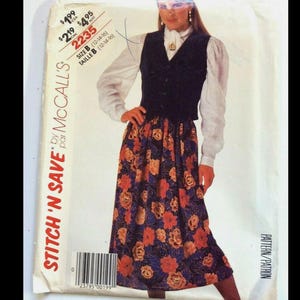 May include: A vintage McCall's sewing pattern featuring a woman's outfit. The outfit includes a dark vest, a white blouse, and a long skirt with a floral pattern in shades of orange, blue, and purple. The pattern size is B (12-14-16).