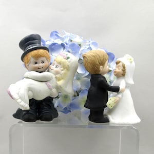 May include: Two ceramic wedding cake toppers featuring a pair in formal attire. One figure wears a black top hat and carries a partner in a white dress. The second figure wears a black suit and stands facing a partner in a white dress and veil.
