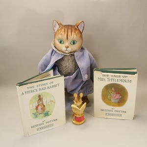 May include: A plush cat doll wearing a blue outfit stands with two vintage Beatrix Potter books and a small figurine. The books are titled "The Story of a Fierce Bad Rabbit" and "The Tale of Mrs. Tittlemouse."