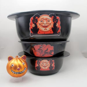 May include: A stack of three black Halloween bowls with a wide rim. Each bowl has a different vintage-style sticker. One features a smiling moon with black cats, another a spooky carriage, and the last one the same moon and cats.