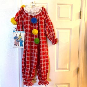 May include: A red and white plaid clown jumpsuit featuring a white ruffled collar, long sleeves with gathered cuffs, and three large pom-poms in blue, yellow, and green down the front. A Simplicity sewing pattern envelope is clipped to the side.