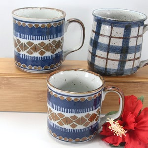 May include: Three ceramic mugs with handles. Two mugs have a blue and brown geometric pattern, and one has a blue and white plaid design. The mugs are on a wooden box with a red flower in the foreground.