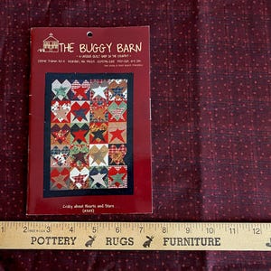May include: A red and white fabric with a pattern of small dots and a quilt pattern book. The book is titled "The Buggy Barn" and features a quilt design with hearts and stars. A wooden ruler is at the bottom.