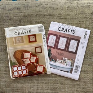May include: Two McCall's Crafts sewing pattern envelopes. The left envelope, number 3554, shows red and white pillows in a wicker chair. The right envelope, number M6001, displays three framed holiday-themed cross-stitch designs on a table.