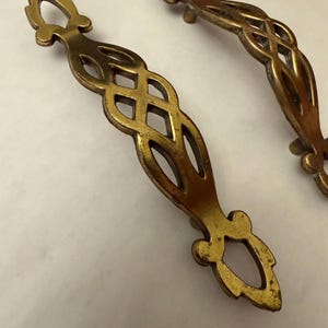 May include: Two antique brass cabinet pulls with an openwork design. The pulls have a decorative, elongated shape with a central diamond pattern and ornate ends. The brass has a warm, aged patina.