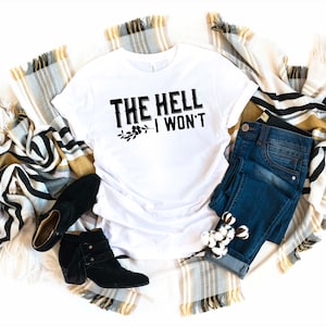 May include: White t-shirt with black text that reads "THE HELL I WON'T" with a small floral design below the text. The shirt is laid out on a white surface with a blanket and jeans.