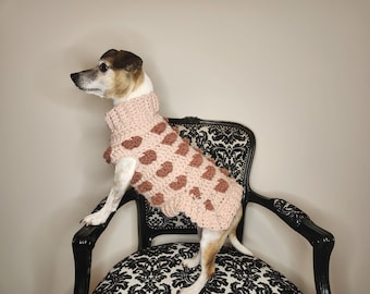May include: A small white dog wearing a pink and brown crocheted sweater with a heart pattern. The dog is sitting on a black chair with a black and white patterned upholstery.