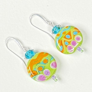 May include: A pair of handmade earrings featuring large, round, colorful glass beads. The beads are primarily lime green with orange, blue, and pink abstract designs. Each earring has a silver-tone ear wire and a small turquoise bead.