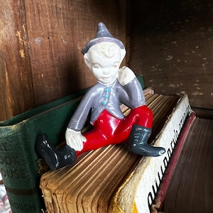 May include: A gray and red figurine of a gnome wearing a pointed hat and sitting on a stack of books. The gnome has a white face with blue eyes and a red nose.
