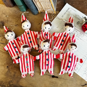 May include: Seven handmade felt Christmas ornaments shaped like people wearing red and white striped outfits with white lace trim. The ornaments have brown hair and painted faces.