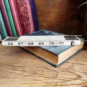 May include: A white wooden folding ruler with black markings and red numbers. The ruler is open to show the numbers 66 to 71 inches.