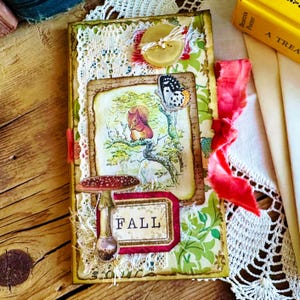 May include: A handmade journal with a fall theme. The cover features a red squirrel, a mushroom, a butterfly, and a tag that says "FALL". The journal is decorated with lace, fabric, and other embellishments.