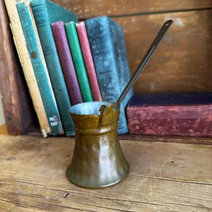 May include: A vintage copper coffee pot with a long handle. The pot has a hammered finish and is sitting on a wooden surface.