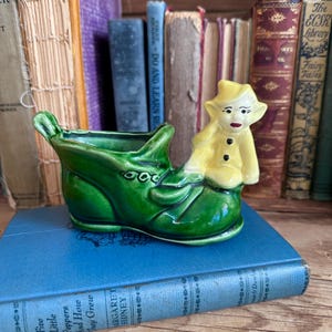 May include: A ceramic planter shaped like a green boot with a yellow elf figurine. The boot is glossy green with decorative details. The elf wears a yellow outfit with black buttons and a red mouth. The planter sits on a blue book.