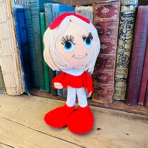May include: A handmade doll with a light skin tone, blonde hair, and blue eyes. The doll wears a red outfit with white pants and red shoes. The doll is standing in front of a shelf of vintage books.