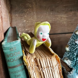May include: A yellow ceramic figurine of a person with a green hat lying on top of a stack of old books. The figurine has red lips and green eyes.