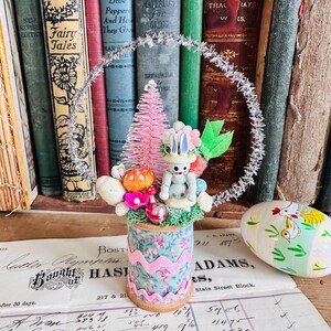 May include: A whimsical Easter decoration featuring a miniature bunny figurine with a floral hat, nestled amidst colorful flowers and a pink bottle brush tree. The arrangement sits atop a spool decorated with floral fabric and pink ribbon, with an Easter egg nearby.