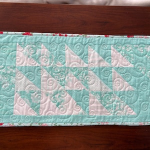 May include: A teal and white quilted table runner with a floral print border. The quilt is made of fabric squares with a swirling pattern.