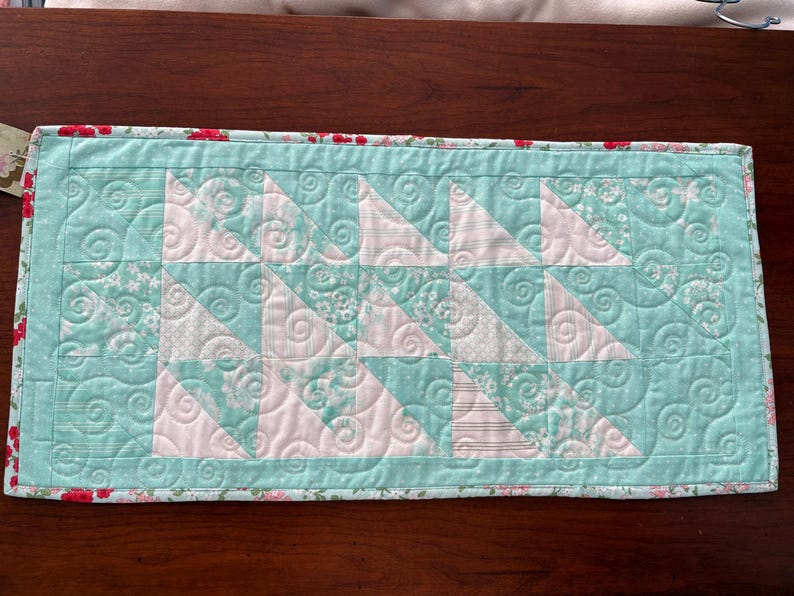 May include: A teal and white quilted table runner with a floral print border. The quilt is made of fabric squares with a swirling pattern.