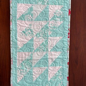 May include: A quilted table runner with a pink and green patchwork design. The runner has a floral print border and a swirl quilting pattern.
