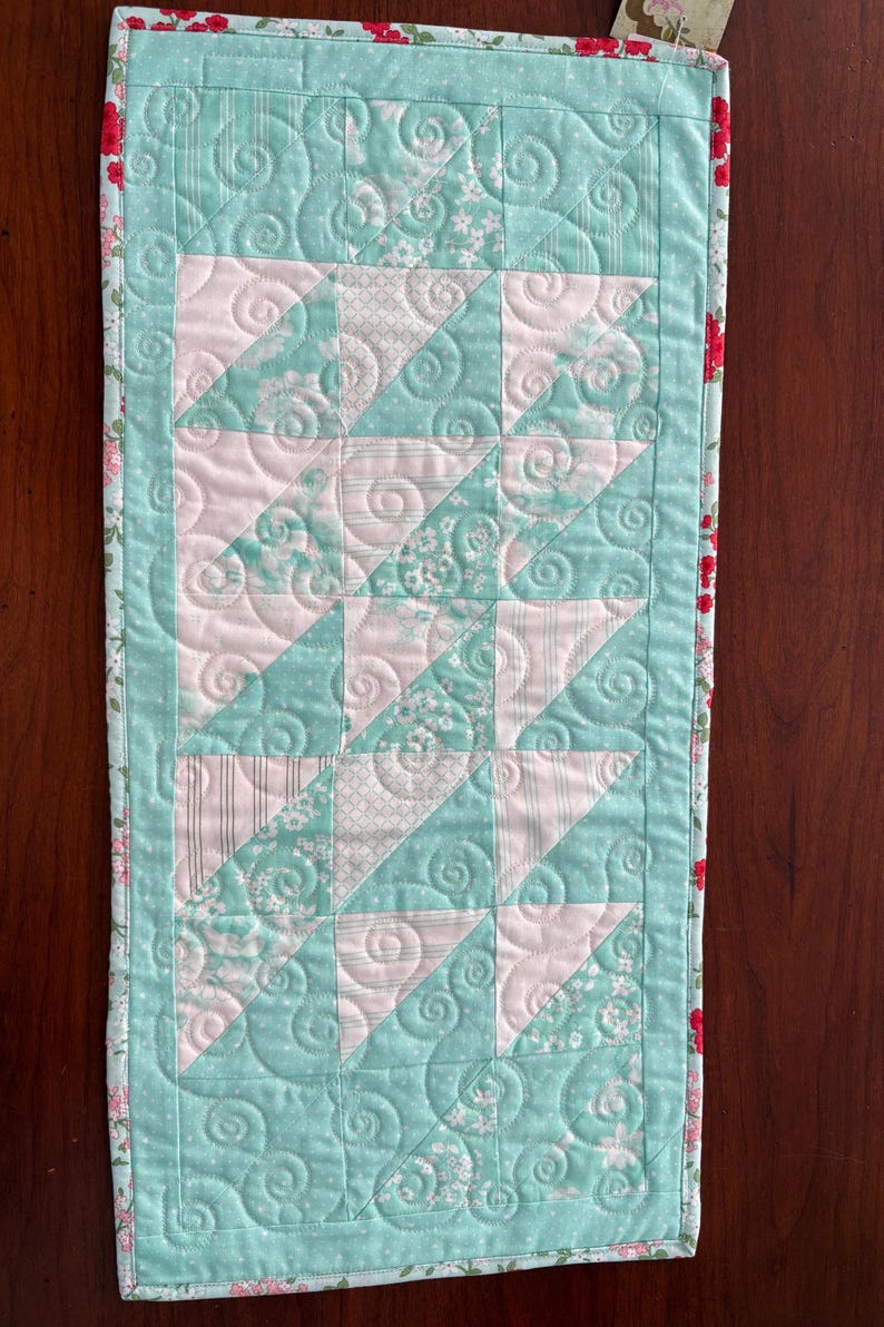 May include: A quilted table runner with a pink and green patchwork design. The runner has a floral print border and a swirl quilting pattern.