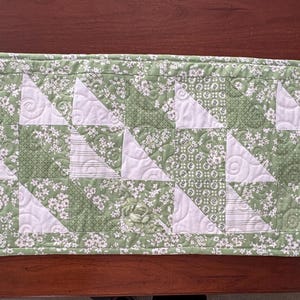May include: A green and white quilted table runner with a floral pattern. The runner is made of fabric squares sewn together in a geometric pattern.